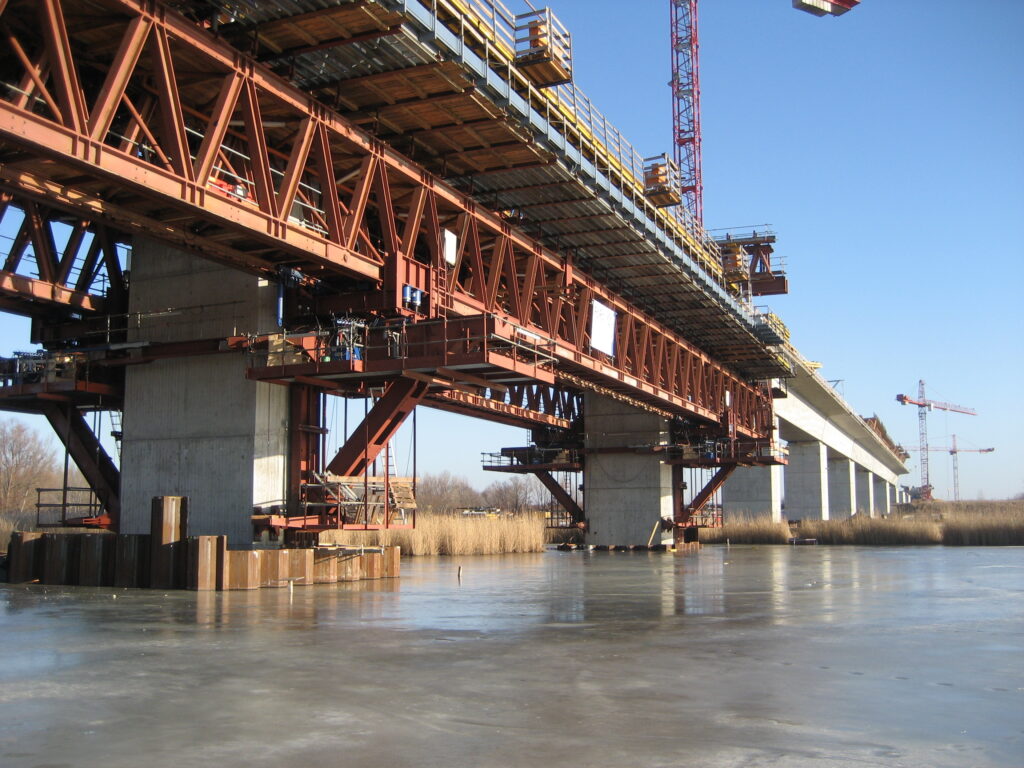 Brücke im Bau mit Stahlträgern und Kränen, die über einen gefrorenen Gewässerbereich führt.