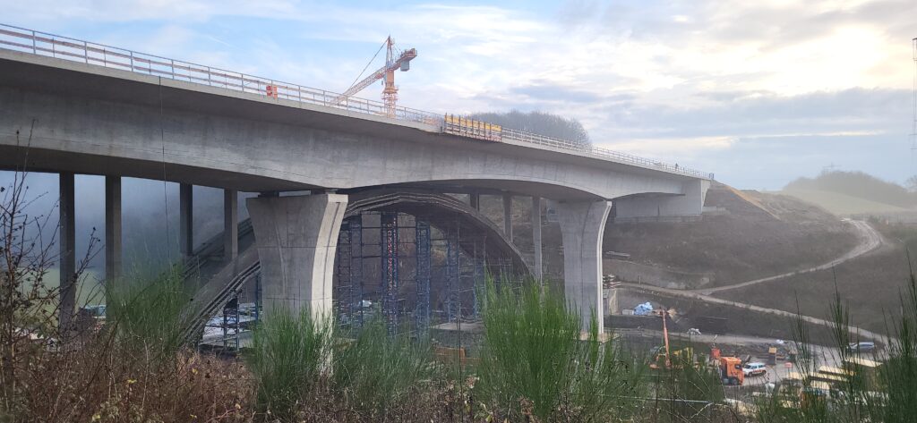 Brücke in der Bauphase mit sichtbaren Betonstrukturen und Baugeräten im Hintergrund.