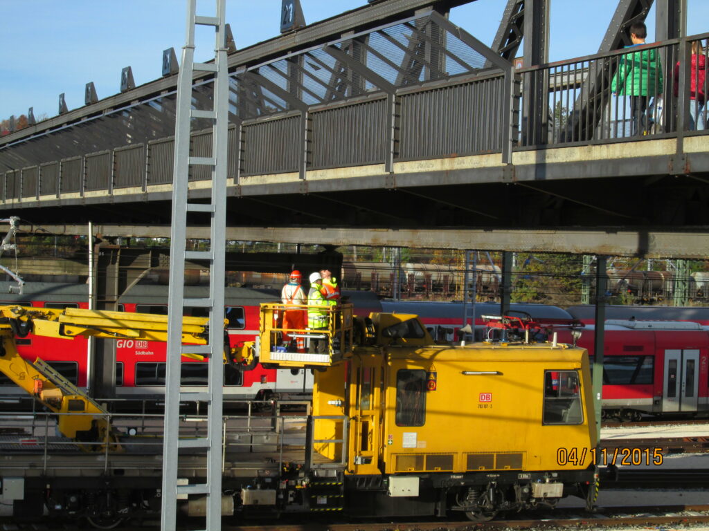 Gelbes Gleisbaufahrzeug mit Arbeitern auf einer Brücke über einem Bahnhof.