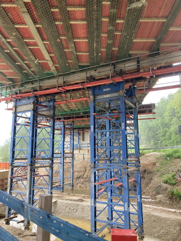 Baugerüst mit blauen Stützen und rotem Schalungsdeckel unter einer Brücke in einem Baustellenbereich.