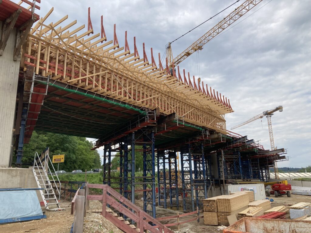 Baugerüst mit Holzschalung für eine Brücke, umgeben von Baukränen und Baumaterialien.