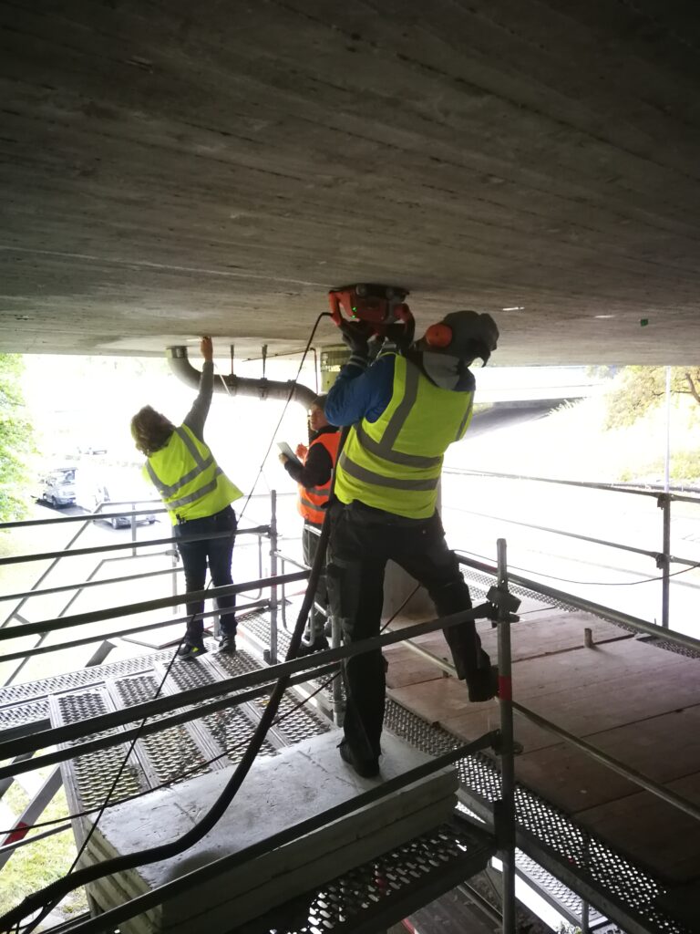 Arbeiter in Sicherheitswesten, die an einer Betonunterseite mit Werkzeugen arbeiten.