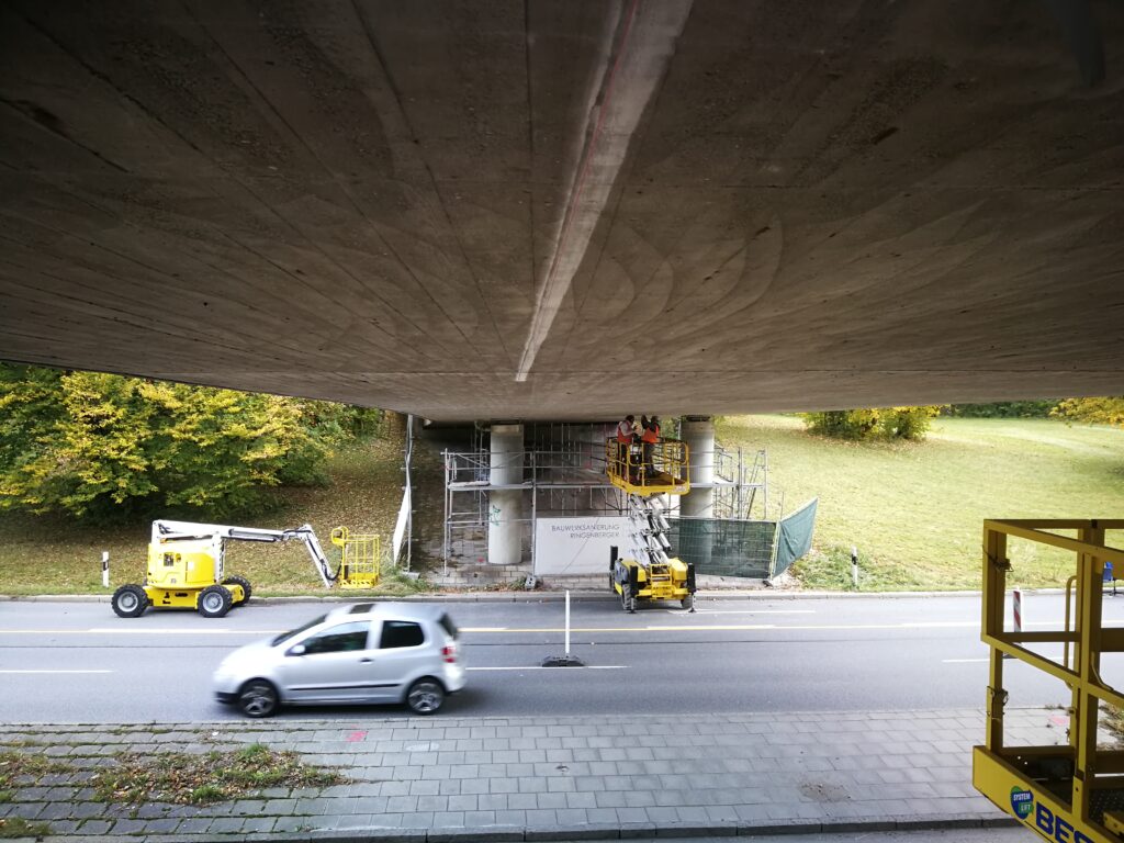 Blick auf eine Baustelle unter einer Brücke mit Baugeräten und einem Auto auf der Straße.