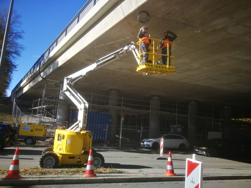 Zwei Arbeiter in einem Arbeitskorb, der an einem Kran unter einer Brücke hängt, während sie an einem Kabel arbeiten.
