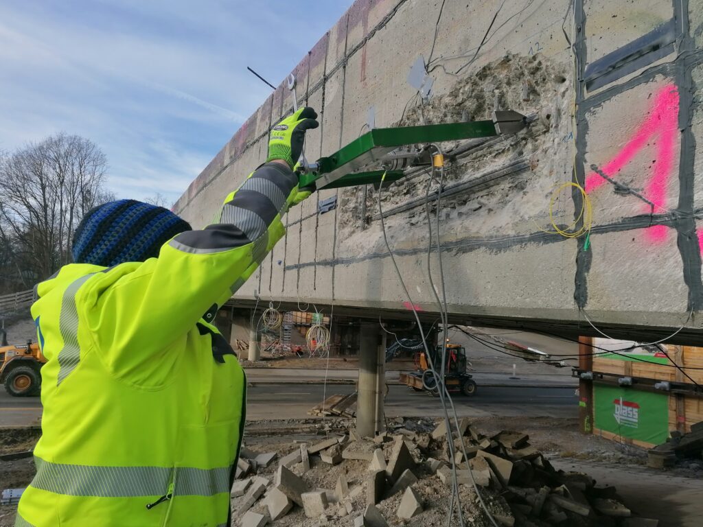 Ein Ingenieur in einer leuchtend gelben Jacke untersucht eine Betonstruktur mit einem Messgerät auf einer Baustelle.