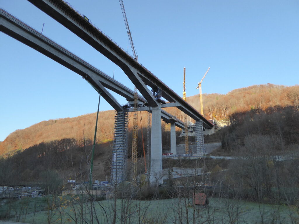 Bau einer Brücke mit mehreren Pfeilern und Kränen im Hintergrund, umgeben von Hügeln.