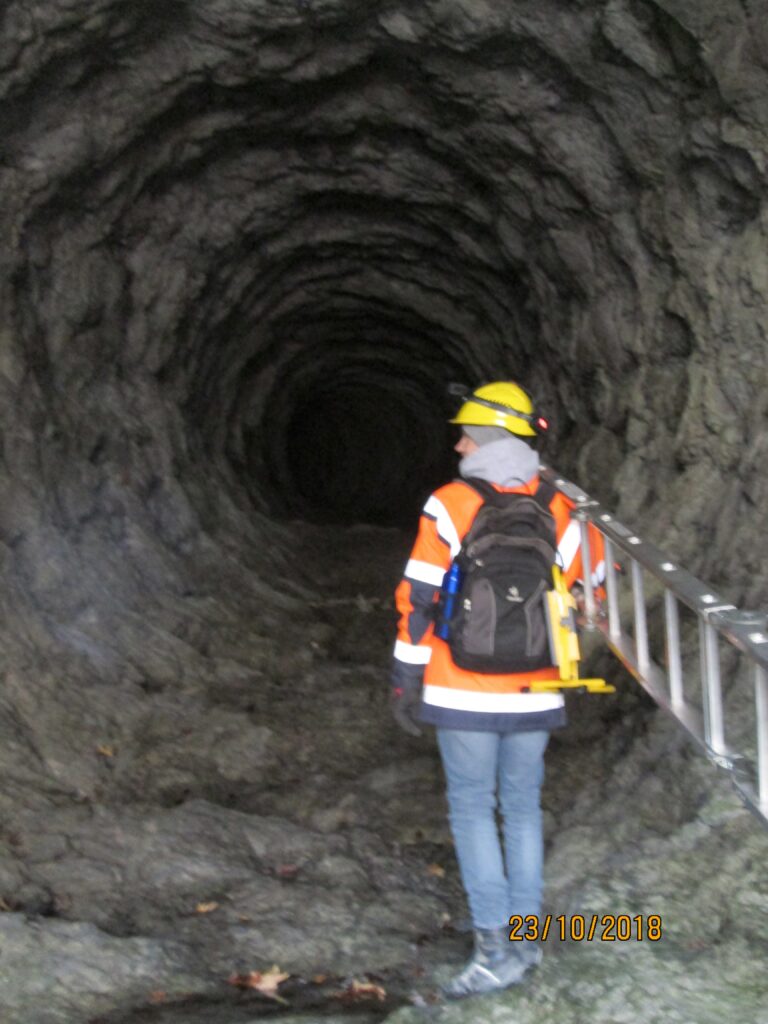 Person in orange Sicherheitsjacke und Helm steht in einem Tunnel mit einer Leiter im Hintergrund.